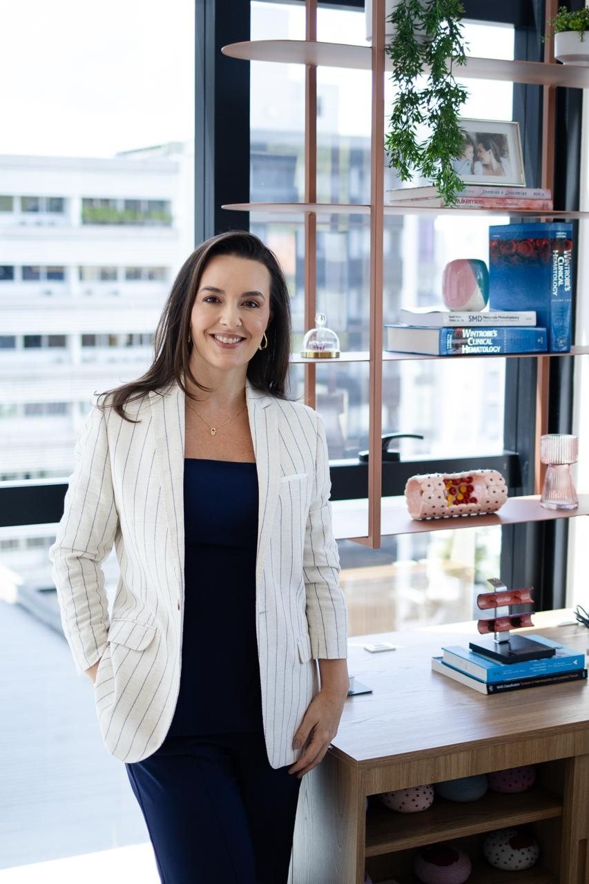 Dra. Andrezza Medina no consultório, ao lado da biblioteca de hematologia